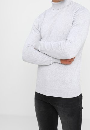 Osoba ubrana w jasnoszary golf i czarne spodnie, ze skrzyżowanymi ramionami na jednolitym białym tle.