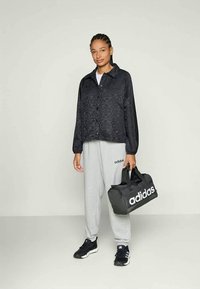 Svart mönstrad jacka med knäppning, grå träningsbyxor, svarta sneakers och en svart väska med vit Adidas-logotyp. Avslappnad outfit.
