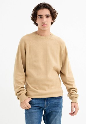 Suéter de punto beige con cuello redondo, puños acanalados y un ajuste relajado. Usado sobre jeans azules, con una textura suave y mangas raglán.