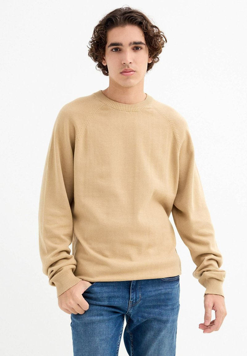 Suéter de punto beige con cuello redondo, puños acanalados y un ajuste relajado. Usado sobre jeans azules, con una textura suave y mangas raglán.