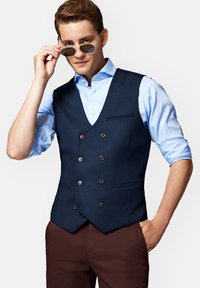 Granatowa kamizelka z teksturowanej tkaniny, dwurzędowa, z metalowymi guzikami, zestawiona z jasnoniebieską koszulą i okularami przeciwsłonecznymi.