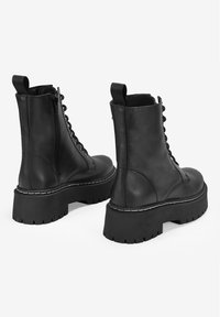 Botas de tobillo de cuero negro con suela de goma gruesa, cordones en la parte delantera y detalle de cremallera en el costado; tacón plano y parte superior texturizada.
