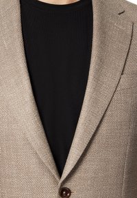 Jasnobrązowa marynarka o fakturze z wzorem w jodełkę, z pojedynczym zapięciem na guzik oraz czarną koszulką z okrągłym dekoltem pod spodem.