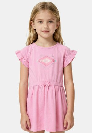 Jeune fille aux longs cheveux blonds portant une robe rose à manches volantées et une ceinture avec un nœud, debout devant un fond uni.