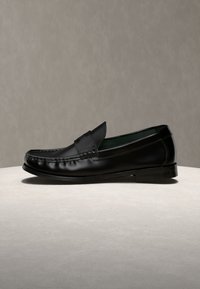 Μαύρο δερμάτινο penny loafer με ραμμένες λεπτομέρειες και χαμηλό τακούνι, εμφανίζεται σε φωτεινή επιφάνεια πάνω σε ουδέτερο φόντο.