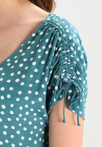 Blusa de color azul verdoso con lunares blancos, detalles en las mangas fruncidas y acentos con cordón. Tejido suave y texturizado con un ajuste relajado.