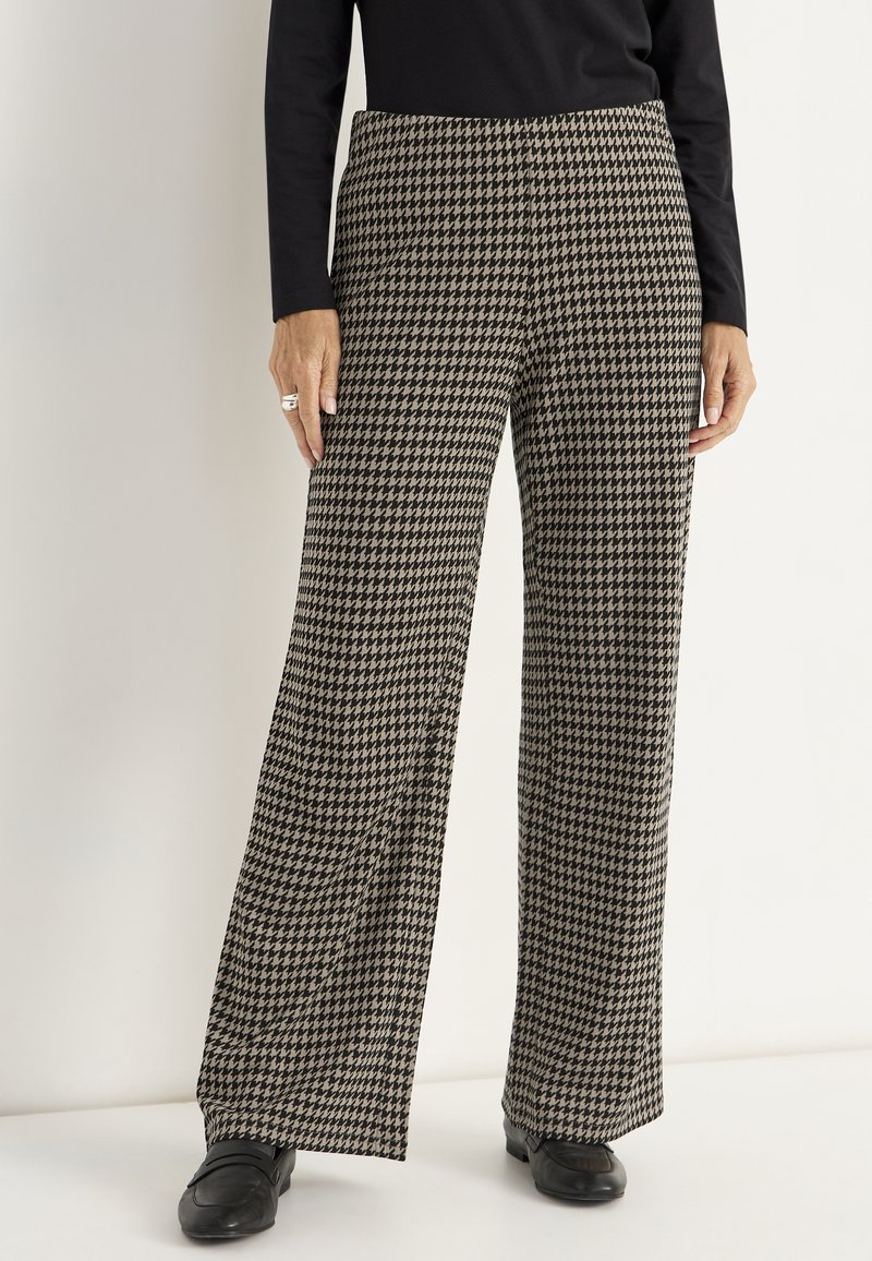 Vrouw in zwart en beige houndstooth wijde broek, zwart langemouwen top en zwarte instappers, staande tegen een witte achtergrond.