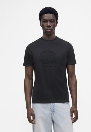 Giovane uomo con pelle scura che indossa una maglietta nera con grafica e jeans azzurri, in piedi davanti a uno sfondo bianco uniforme.