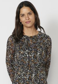 Dunkelbluse mit floralem Muster, die einen runden Ausschnitt, lange Ärmel und ein Schlüsselaugen-Detail vorne aufweist. Die Muster umfassen grüne, lila und braune Blumen.