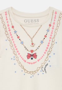 Tricou crem cu modele strălucitoare roz și albastre, inclusiv lanțuri, stele, o fundă și textul „Guess”. Textură netedă a țesăturii.