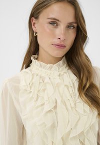 Roomwitte blouse met ruches, hoge kraag en knoopdetails. Transparante stof met een gestructureerde uitstraling, met gelaagde, vloeiende ruches aan de voorkant.