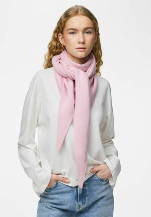 Nuori nainen, jolla on kiharat vaaleat hiukset, pukeutunut valkoiseen pitkähihaiseen paitaan, sinisiin farkkuihin ja kaulassaan vaaleanpunainen neulottu huivi.