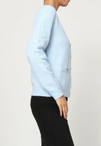Cardigan bleu clair en matériau doux, avec des boutons dorés et des poches, des poignets et un ourlet côtelés, porté avec un pantalon noir.