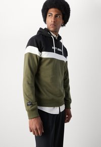 Oliven grønn hettegenser med svart og hvit horisontale striper, med et frontlogo, justerbare snorer og ribbestrikkede mansjetter.