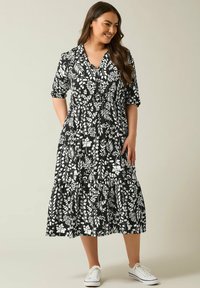 Schwarz-blumiges Midikleid mit kurzen Ärmeln, einem V-Ausschnitt und einem gestuften Rock. Mit weißen botanischen Drucken und seitlichen Taschen. Trägt man mit weißen Sneakers.