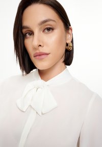 Blusa blanca con un lazo en el cuello, textura translúcida y caída suave. Pendientes de aro dorados realzan el look. Tez de piel suave.
