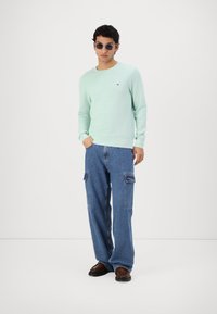 Pull à manches longues vert menthe, texture côtelée, logo sur la poitrine. Pantalons cargo bleus avec poches, coupe décontractée. Chaussures marron foncé avec une finition lisse.
