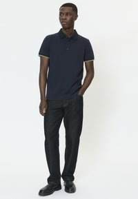 Polo azul marino con puños de rayas en contraste de color, ajuste entallado, bolsillo en el pecho, combinado con vaqueros de pierna recta en denim oscuro y zapatos negros.