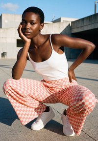 Young woman squatting outdoors wearing a white tank top, pink and white checkered pants, and white sneakers, resting her face on her hand.