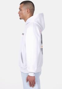 Haut de hoodie blanc avec une poche kangourou, un design graphique sur le côté, une capuche spacieuse et des poignets côtelés. Le matériau semble doux et texturé.