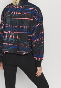 Osoba oblečená v černém trikotu s dlouhým rukávem, na kterém je abstraktní textový design v růžové a modré, a v černých legínách, stojící s zády k fotoaparátu.