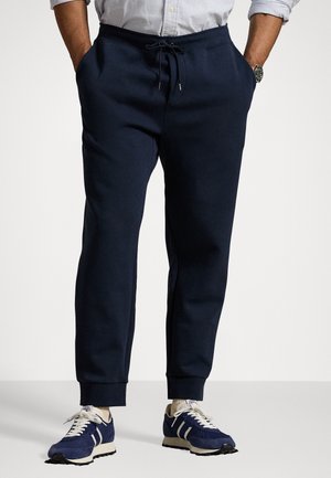 Mann trägt marineblaue Jogginghose mit Kordelzug und weißen Sneakers, Hände in den Taschen, steht vor einem einfarbigen Hintergrund.