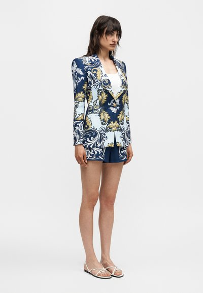 Γυναίκα με μπλε σκούρο και χρυσό μπλε ζακέτα με μπαρόκ σχέδιο με αντίστοιχο σορτς και λευκό τοπ, φοράει λευκά σανδάλια και στέκεται μπροστά σε απλό φόντο.