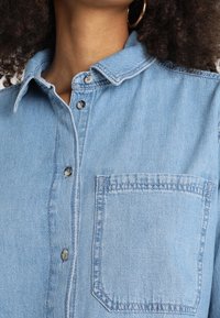 Ljust blå denimskjorta med krage som knäpps, bröstficka och synliga sömmar. Tyget har en strukturerad yta.