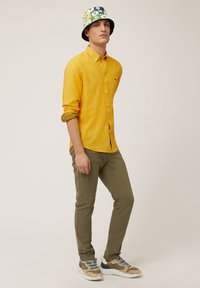 Camisa amarilla de botones, pantalones verde oliva y zapatillas beiges con acentos azules. El modelo lleva un sombrero de cubo con estampado floral.