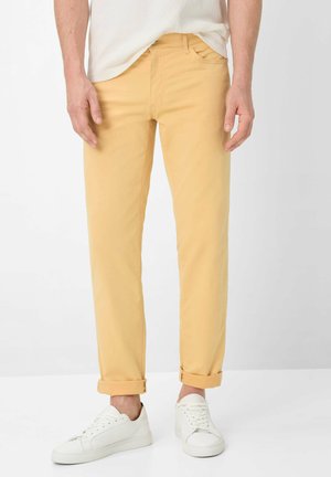 Mann trägt hochgekrempelte senfgelbe Hosen und weiße Sneakers, steht vor einem schlichten weißen Hintergrund.