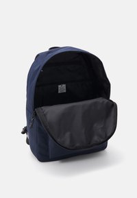 Sac à dos bleu marine avec un extérieur en tissu solide. Rabat ouvert révélant un intérieur noir avec un compartiment plat et une étiquette sur le mur arrière.