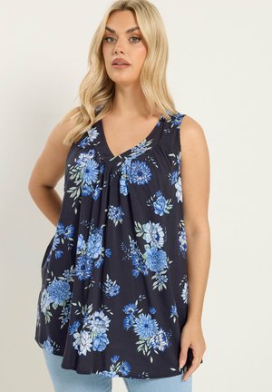 Frau trägt ein ärmelloses, marineblaues Top mit blauem und weißem Blumenmuster, steht mit einer Hand in der Hüfte vor einfarbigem Hintergrund.