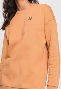 Sudadera de cuello redondo naranja hecha de un tejido suave. Presenta un logotipo bordado amarillo en el pecho y mangas de corte relajado.