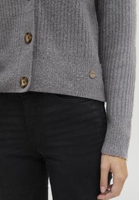 Grå ribbestrikket cardigan med brune knapper, som har en myk tekstur og fitted silhuett, sammen med svarte bukser.