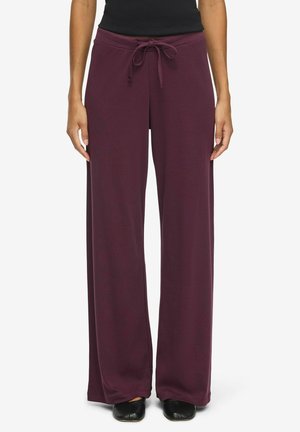 Personne portant un pantalon large ample violet foncé avec une taille à cordon et des chaussures noires, debout, les bras détendus le long du corps.