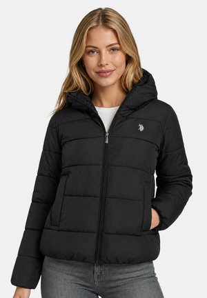 Young woman with long blonde hair wearing a black puffer jacket with a hood, standing with one hand in pocket and smiling slightly.