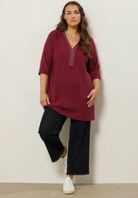 Tunic bordeaux con scollo a V e borchie decorative lungo il colletto, abbinata a pantaloni in denim scuri a gamba larga e sneakers bianche.
