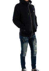Felpa nera con zip e tasca frontale, hardware argentato e logo sul petto. Indossata con jeans blu strappati e sneakers a fantasia.