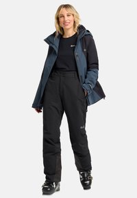 Waterproof donkerblauwe en zwarte skijas gecombineerd met zwarte geïsoleerde broek en skischoenen. Voorzien van ritszakken en verstelbare manchetten.