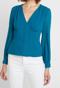 Türkisfarbene Bluse mit langen Ärmeln, tiefem V-Ausschnitt, gerafftem Detail unter der Brust, Peplum-Taille und Knopfverschluss vorne. Glattes Material.
