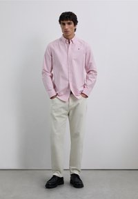 Lyserød skjorte med knapper og lodrette striber, kombineret med cremefarvede bukser og sorte sko. Skjorten har krave og logo på brystet.