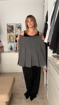 Jupe plissée grise au toucher lisse, tenue par une personne en haut noir. Portant un portant de vêtements et de la décoration en arrière-plan.