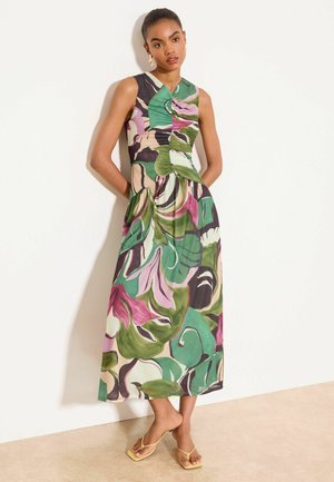 Kvinde stående mod en væg iført en ærmeløs, V-hals, ankel-lang kjole med grønne, pink og lilla abstrakte bladmønstre og gule sandaler.