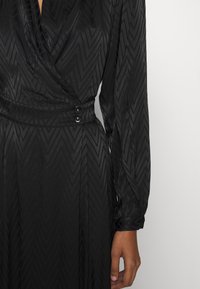 Mustavärinen kietaisumekko, jossa on pitkät hihat, valmistettu sileästä kankaasta, jossa on hienovarainen chevron-kuvio. Mukana on kaksi mustaa nappia vyötär öllä.