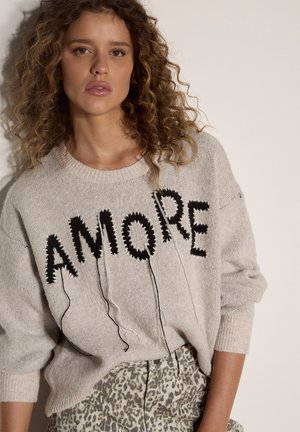 Donna con capelli ricci indossa un maglione beige con lettere "AMORE" nere ricamate e pantaloni a vita alta con motivo contro un muro semplice.