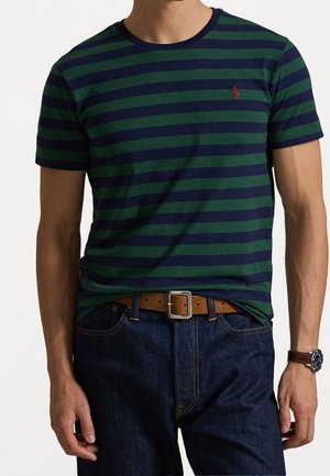 Mand iført navy- og grønstribet kortærmet skjorte, mørkeblå jeans, brun bælte med sølvspænde og armbåndsur.
