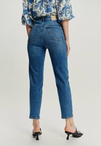 Mujer vestida con vaqueros azul de cintura alta, una blusa blanca con estampado floral azul y mule negros de tacón, vista desde atrás.