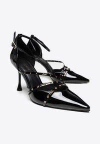Scarpe con tacco alto in vernice nera con punte appuntite, cinturini alla caviglia incrociati e borchie decorative dorate sui cinturini.