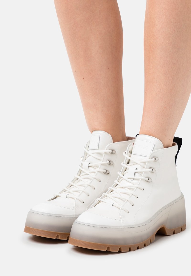 Bottes blanches montantes à lacets avec semelles épaisses translucides et semelle marron portées sur jambes nues sur un fond uni.