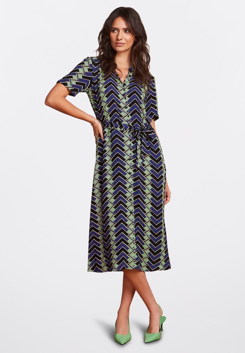 Dress features a navy background with green zigzag patterns, short sleeves, button front, and a tie belt at the waist. Green heels complement.